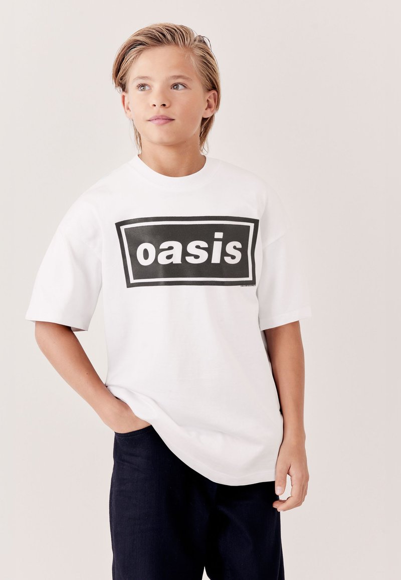 Junge mit kurzem blondem Haar, trägt ein weißes übergroßes T-Shirt mit dem Wort "oasis" in einem schwarzen Rechteck, die Hände in den Taschen dunkler Hosen.