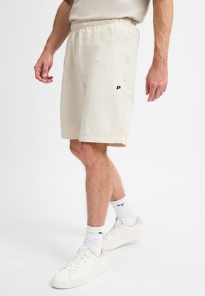Man draagt off-white korte broek met elastische tailleband, witte sokken en witte sneakers, staand op een effen witte achtergrond.