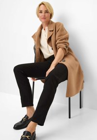 Kameelwolblendenjas met oversized pasvorm, opgerolde mouwen en zwarte knopen; gecombineerd met zwarte slim-fit broek en zwarte loafers met gouden accenten.