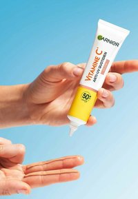Tube de crème solaire avec un design blanc et jaune, étiqueté "Vitamine C," avec un bec verseur distribuant un produit beige clair sur deux doigts devant un fond bleu.