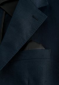 Revers de blazer bleu marine foncé avec un mouchoir de poche noir glissé dans la poche poitrine, montrant la fine texture du tissu et les détails de couture.