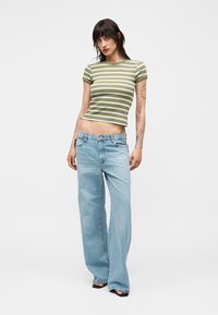 Femme portant un t-shirt court rayé vert et blanc, un jean bleu clair à jambes larges et des chaussures noires ouvertes, debout devant un fond uni.