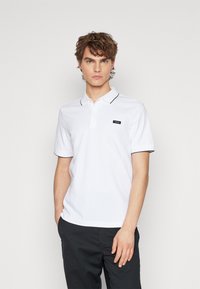 Polo bianco con un marchio nero, colletto e bordi delle maniche neri a contrasto, realizzato in un tessuto liscio e traspirante.