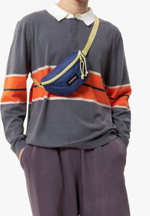 Modrá ledvinka se žlutými akcenty, zakřivený tvar, přední zip a logo. Nosit na šedém pruhovaném tričku s oranžovými a zelenými detaily.