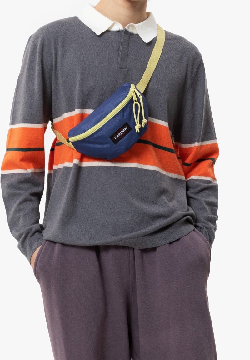 Sininen vyölaukku, jossa on keltaisia yksityiskohtia, kaareva muoto, edessä vetoketju ja logo. Päällä harmaa raidallinen paita, jossa on oransseja ja vihreitä yksityiskohtia.