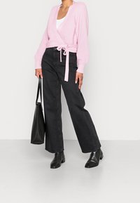 Suéter cruzado de color rosa claro con textura acanalada y un lazo en la cintura, combinado con jeans negros de pierna ancha y botas negras con textura y tacón. Sostiene un bolso negro.