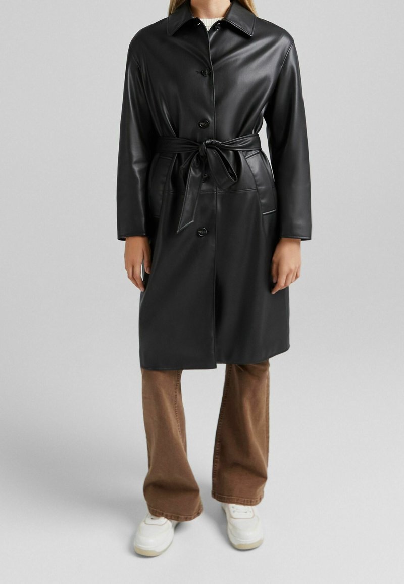 Personne portant un manteau en cuir noir ceinturé, un pantalon marron et des baskets blanches sur un fond gris clair uni.