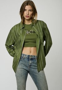 Camicia verde con bottoni e linee ondulate bianche, indossata sopra un crop top verde e jeans di denim blu. Presenta una tasca con patch e una vestibilità rilassata.