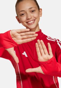 Rode sporttop met een hoge kraag, lange mouwen en duimgaten. Bevat witte accenten en een logo op de borst. Gladde textuur.