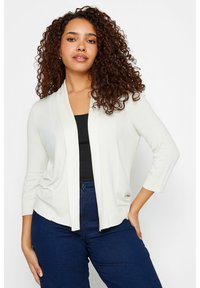 Cárdigan de color crema claro con cuello chal, diseño de apertura frontal, mangas tres cuartos y una textura de punto suave, combinado con jeans azul oscuro.