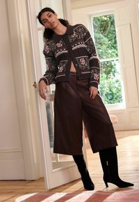 Gehaakte bloemen cardigan in donkerbruin met roze accenten, gecombineerd met hoge bruine faux leren culottes en zwarte laarzen met hoge hakken tot de knie.