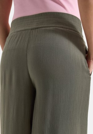 Nærbillede af person iført olivengrønne teksturerede viddebukser og en indstukket lyserød top, hånd hvilende i lommen.