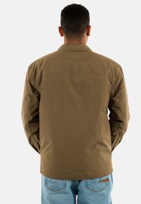 Giacca leggera khaki con yoke posteriore, maniche lunghe e vestibilità comoda. Abbinata a jeans in denim blu chiaro, senza motivi o dettagli visibili.