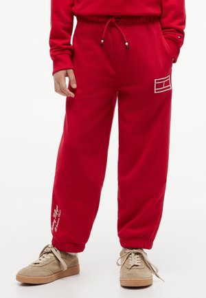 Person med røde joggebukser med snor i livet, hvit brodering på lår og legg, beige joggesko, stående mot en hvit bakgrunn.
