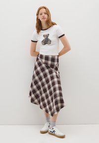 Camiseta blanca de manga corta con estampado de oso, combinada con una falda envolvente a cuadros en rojo y beige, y zapatillas blancas con calcetines grises.
