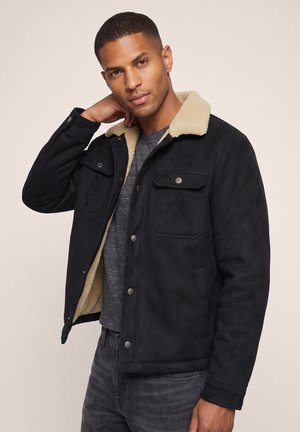 Hombre con chaqueta negra con cuello de borrego beige sobre camisa gris, posando con una mano detrás del cuello sobre un fondo liso.