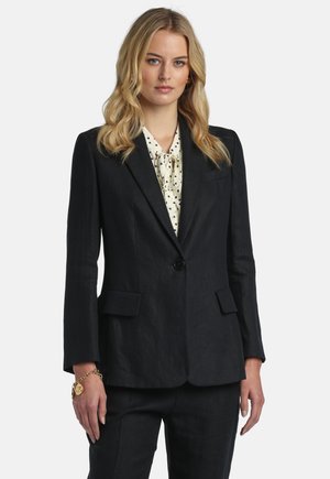 Vrouw met blond haar, gekleed in een zwart op maat gemaakt pak en een witte blouse met zwarte stippen, staand tegen een effen lichte achtergrond.