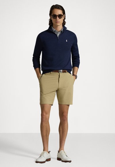 Un pulover cu guler cu fermoar de culoare bleumarin, purtat peste o cămașă cu dungi, asociat cu pantaloni scurți de culoare bej. Pantofi albi și ochelari de soare completează ținuta.