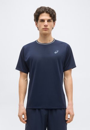 Junger Mann, der nach vorne blickt und ein marineblaues, kurzärmeliges Sportshirt sowie passende Shorts vor einem einfachen Hintergrund trägt.