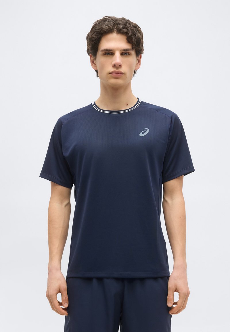 Νέος άνδρας που στέκεται και κοιτάει μπροστά, φορώντας μπλε σκούρο αθλητικό πουκάμισο με κοντά μανίκια και αντίστοιχα σορτς, μπροστά σε απλό φόντο.