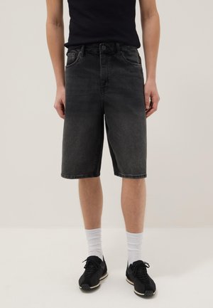 Persona che indossa pantaloncini neri in denim fino al ginocchio, maglietta nera, calzini bianchi e sneaker nere, in piedi su uno sfondo semplice.