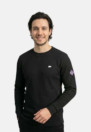 Mann lächelt, trägt einen schwarzen, langärmeligen Waffelstrickpullover mit einem kleinen weißen Bären-Logo auf der Brust und einem Aufnäher am linken Ärmel.