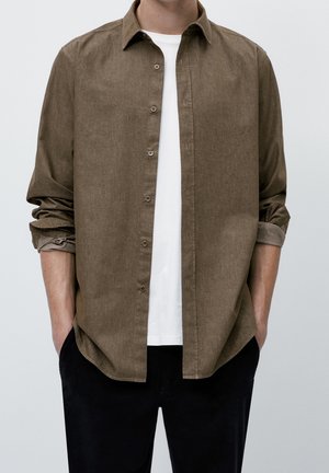 Homme portant une chemise marron à manches longues déboutonnée par-dessus un T-shirt blanc, avec les manches retroussées et les mains dans les poches d'un pantalon noir.