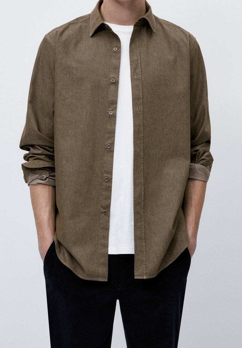 Homme portant une chemise marron à manches longues déboutonnée par-dessus un T-shirt blanc, avec les manches retroussées et les mains dans les poches d'un pantalon noir.