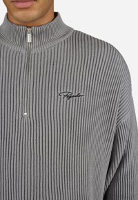 Grå ribbet pullover sweater med høj krave, med en halv lynlås foran og broderet sort logo på brystet. Glat tekstur.