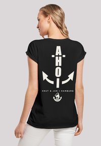 Schwarzes T-Shirt mit großem weißen Text "AHOI", zwei Ankern und "Knut & Jan | Hamburg." Kurze Ärmel und entspannter Schnitt.