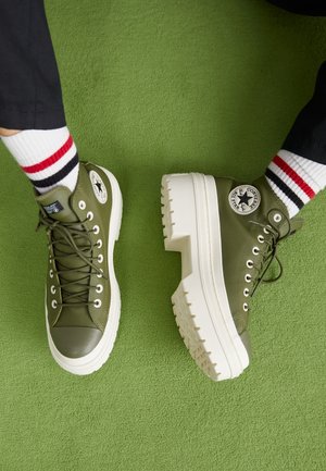 Baskets montantes vertes avec épaisses semelles blanches compensées, portées avec des chaussettes blanches à rayures rouges et noires sur fond de tapis vert.