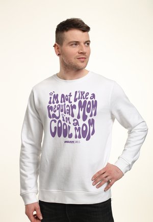 Mann mit kurzen Haaren, der ein weißes Sweatshirt mit lila Schriftzug "Ich bin nicht wie eine normale Mama, ich bin eine coole Mama" aus Mean Girls trägt, Hand in der Hüfte.