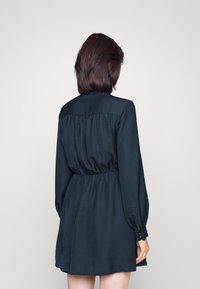 Musta, pitkähihainen mekko, jossa on teksturoidusta kankaasta tehty vyötäröltä kerätty osa ja napitettavat kalvosimet; liehuva helma ylettyy polven yläpuolelle.