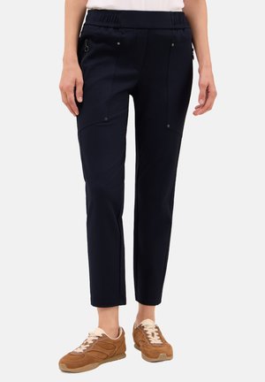 Femme portant un pantalon cropped bleu marine avec des poches zippées sur le côté et des baskets en daim marron, se tenant devant un fond uni.
