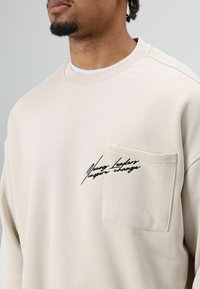 Beige sweatshirt met een ronde hals, voorzien van een borstzak aan de linkerkant en zwarte geborduurde tekst. De stof lijkt zacht met een gladde textuur.