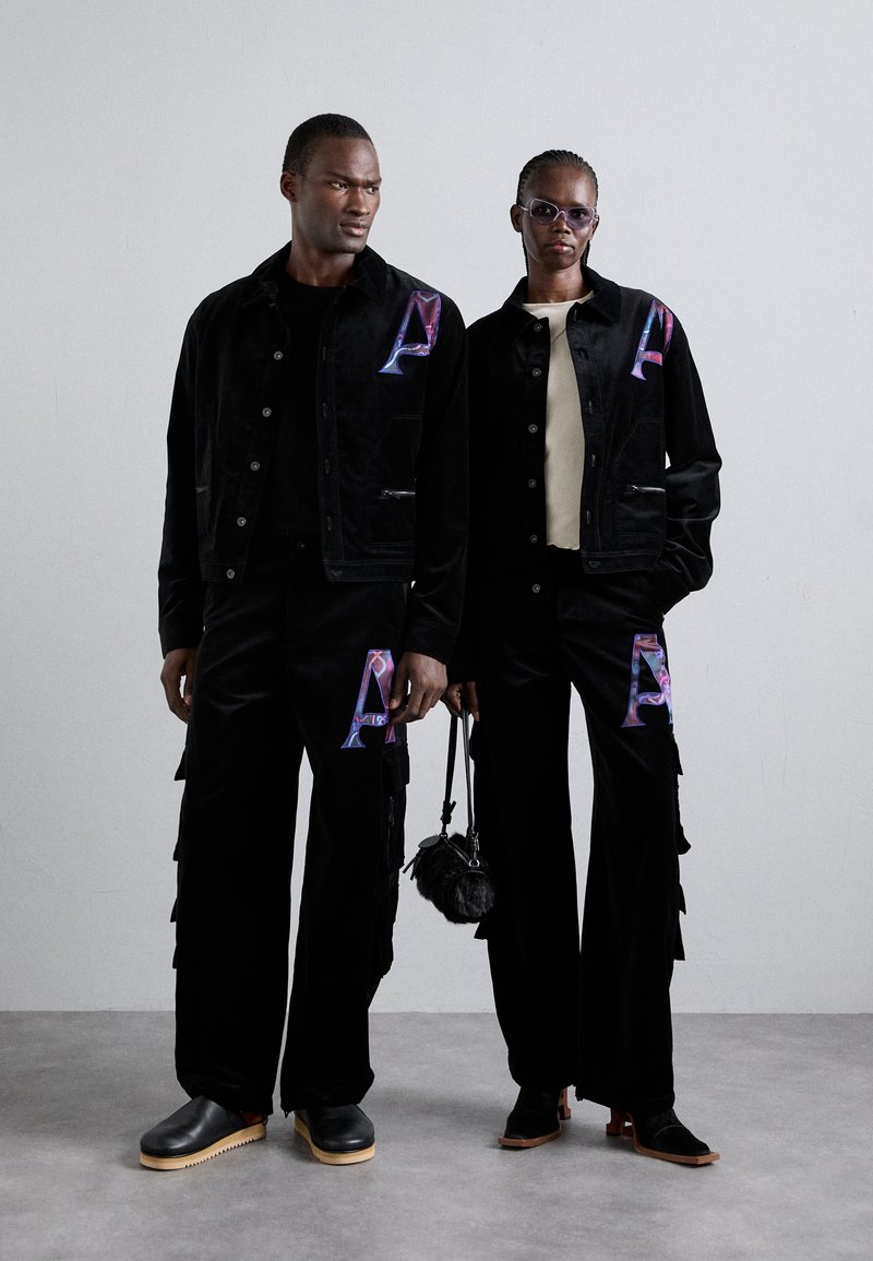 Zwei Models tragen passende schwarze Cordjacken und Hosen mit bunten "A"-Motiven, die mit strukturierten Akzenten versehen sind, ergänzt durch schwarze Schuhe.