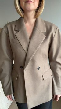 Kvinna iklädd en beige dubbelknäppt kavaj med vida slag och svarta knappar, stående mot en enfärgad bakgrund.