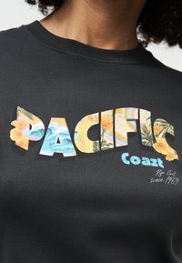 Κοντινό πλάνο μαύρου πουκαμίσου με το κείμενο "PACIFIC Coast" διακοσμημένο με τροπικά λουλούδια και μοτίβα παραλίας, και τη φράση "Rip Curl since 1969" γραμμένη από κάτω.