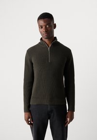 Pulóver de punto acanalado en verde oscuro con cuello de media cremallera y mangas largas. El material tiene una apariencia texturizada, combinado con pantalones negros.