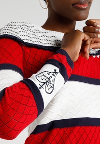 Pull en maille rouge avec un motif texturé, des accents de rayures blanches et marines, et un motif brodé sur la manche.