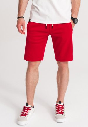 Shorts rouges en tissu doux avec une taille élastique, dotés de cordons de serrage et de poches latérales. Accompagnés de baskets blanches avec des lacets rouges.