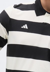 Άνδρας που φοράει μαύρο και λευκό πόλο με οριζόντιες ρίγες, μαύρο γιακά και μικρό λευκό τριγωνικό λογότυπο στο στήθος.