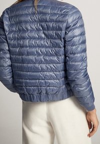 Personne de dos, portant une doudoune matelassée brillante bleu clair et un pantalon blanc sur fond uni.