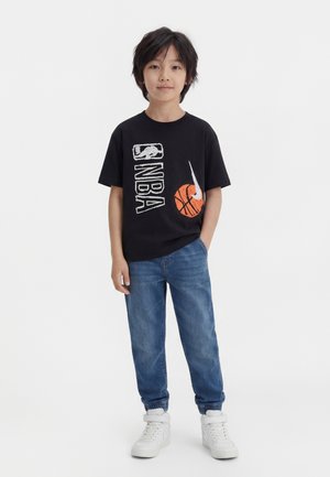 Jeune garçon debout, une main dans la poche, portant un t-shirt noir NBA, un jean bleu et des baskets blanches sur un fond blanc.