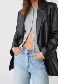 Černý blazer z umělé kůže se strukturovanými rameny, šedý žebrovaný cardigan pod ním a světle modré džíny s vysokým pasem.