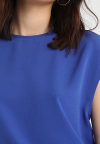 Modrý krátký rukáv top vyrobený z hladké látky, s jednoduchým kulatým výstřihem a čistými liniemi. Žádné viditelné vzory nebo kovové prvky.