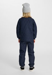 Dziecko ubrane w granatową pikowaną kurtkę i spodnie, szarą czapkę oraz czarne trampki, stojące tyłem na tle jednolitego białego tła.