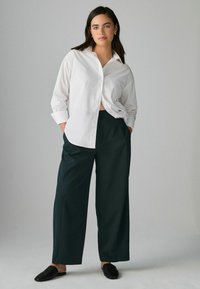 Weißes Hemd mit Knopfleiste, lockere Passform, mit langen Ärmeln. Dunkelgrüne weit geschnittene Hose. Schwarze Slipper vervollständigen das Outfit. Schlichtes, modernes Design.