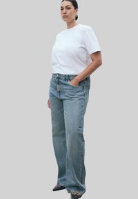T-shirt bianco a maniche corte abbinato a jeans azzurri chiari a vita alta e gamba dritta. Il modello ha le mani nelle tasche, in piedi contro uno sfondo grigio.