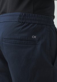 Temno modri trenirki z elastičnim pasom, podrobnostjo na zadnjem žepu in logotipom CK v svetlo sivi barvi na področju žepa. Gladka tekstura.